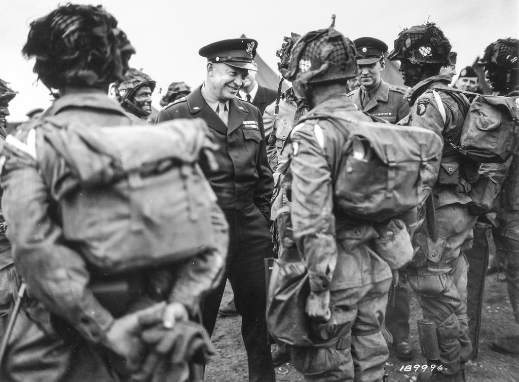 Black and white photo of General Eisenhower in uniform with ribbons and a cap talking to a formation of U.S. airborne troops in complete combat kit to include camouflage paint, ready to move to assault aircraft just prior to D-Day. Black and white photo of General Eisenhower in uniform with ribbons and a cap talking to a formation of U.S. airborne troops in complete combat kit to include camouflage paint, ready to move to assault aircraft just prior to D-Day.