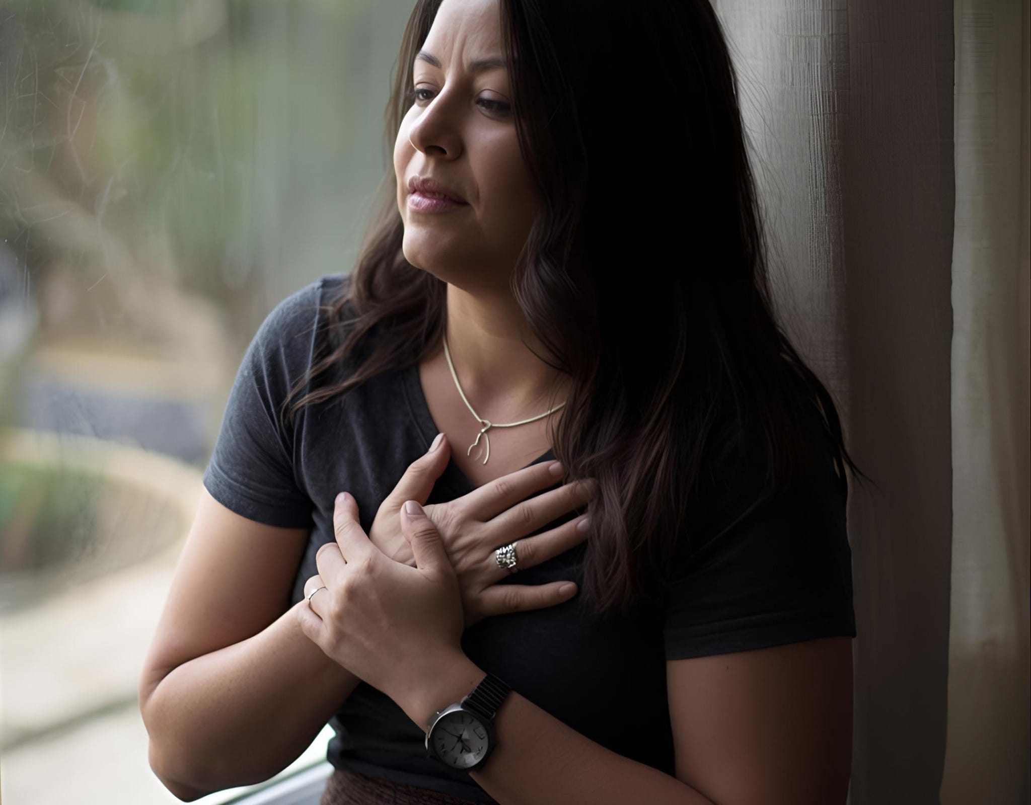 A woman sitting in quiet reflection by a window, hands over her heart, symbolizing emotional healing through mindfulness and presence. The Body Remembers: A Mindful Path to Healing Emotional Pain  Learning to meet what still aches with tenderness and presence