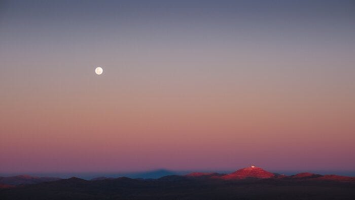 This photograph captures the mesmerising colours of an evening sky. The sky's gradient fades from blue at the top to white, then orange and pink at the bottom. A mountain range is visible at the bottom of the image, with all but one of the mountains already in darkness. The last illuminated one is on the right, with a metallic structure on top. In the middle of the mountain range, a triangular shadow can be seen. A silver full moon high in the sky on the left underlines the timelessness of the image. This photograph captures the mesmerising colours of an evening sky. The sky's gradient fades from blue at the top to white, then orange and pink at the bottom. A mountain range is visible at the bottom of the image, with all but one of the mountains already in darkness. The last illuminated one is on the right, with a metallic structure on top. In the middle of the mountain range, a triangular shadow can be seen. A silver full moon high in the sky on the left underlines the timelessness of the image.