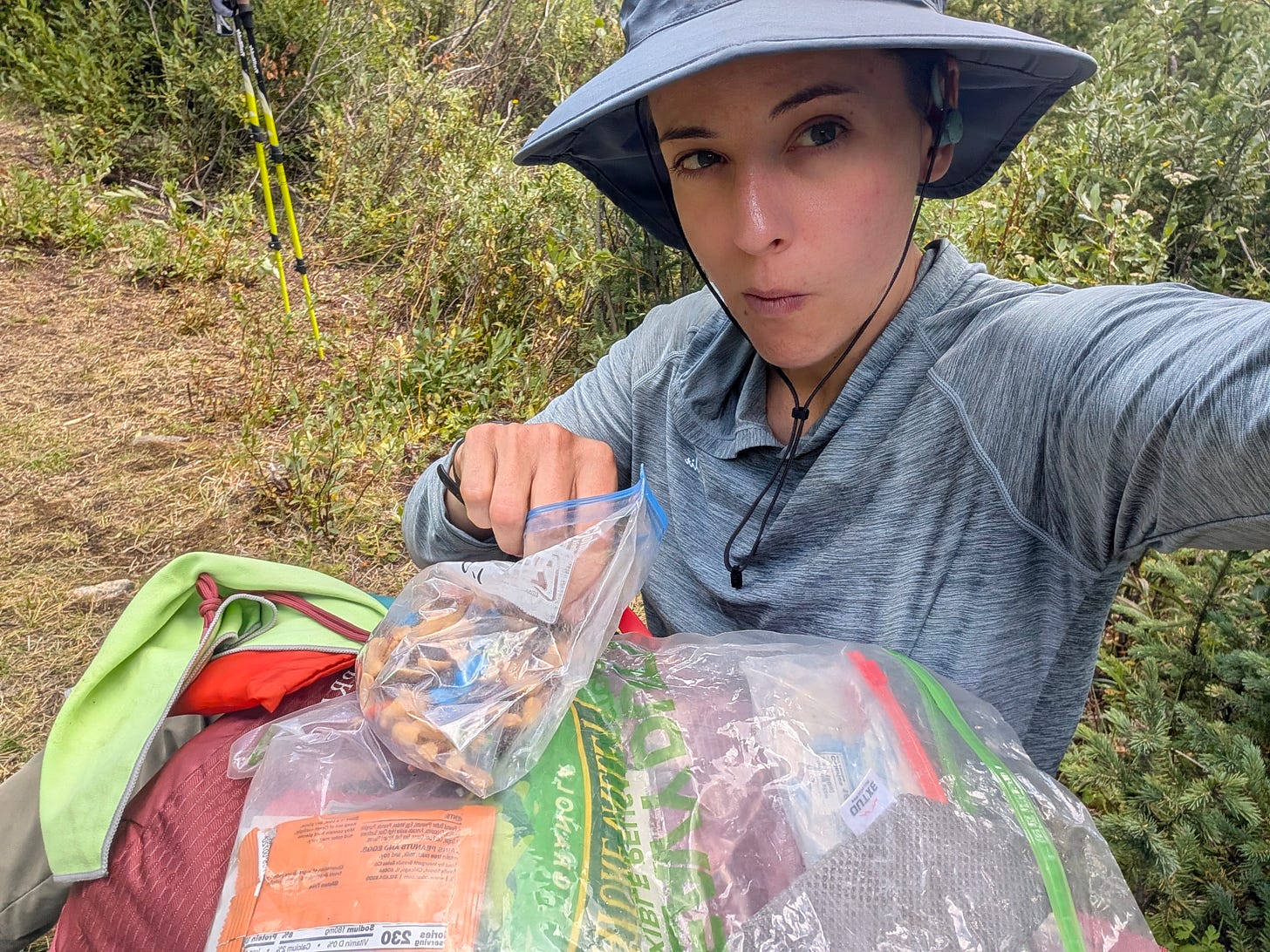 Selfie on trail wearing a wide brim hat, holding an Ursack open and staring down at bags of trail food during a snack break. Selfie on trail wearing a wide brim hat, holding an Ursack open and staring down at bags of trail food during a snack break.