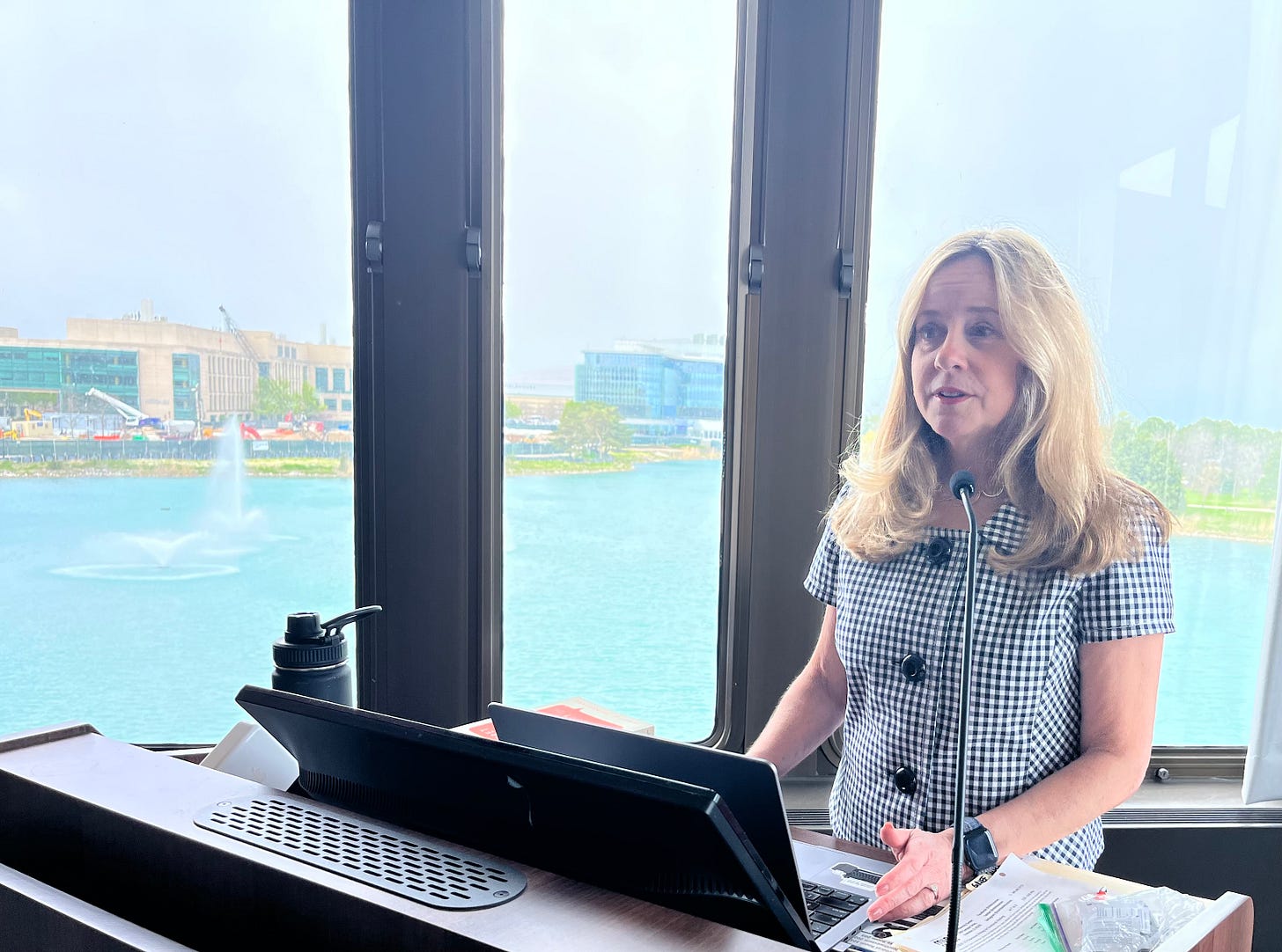 Blonde woman in black and white checkered shirt stands at a podium in front of a window overlooking a blue pond with a water fountain Blonde woman in black and white checkered shirt stands at a podium in front of a window overlooking a blue pond with a water fountain