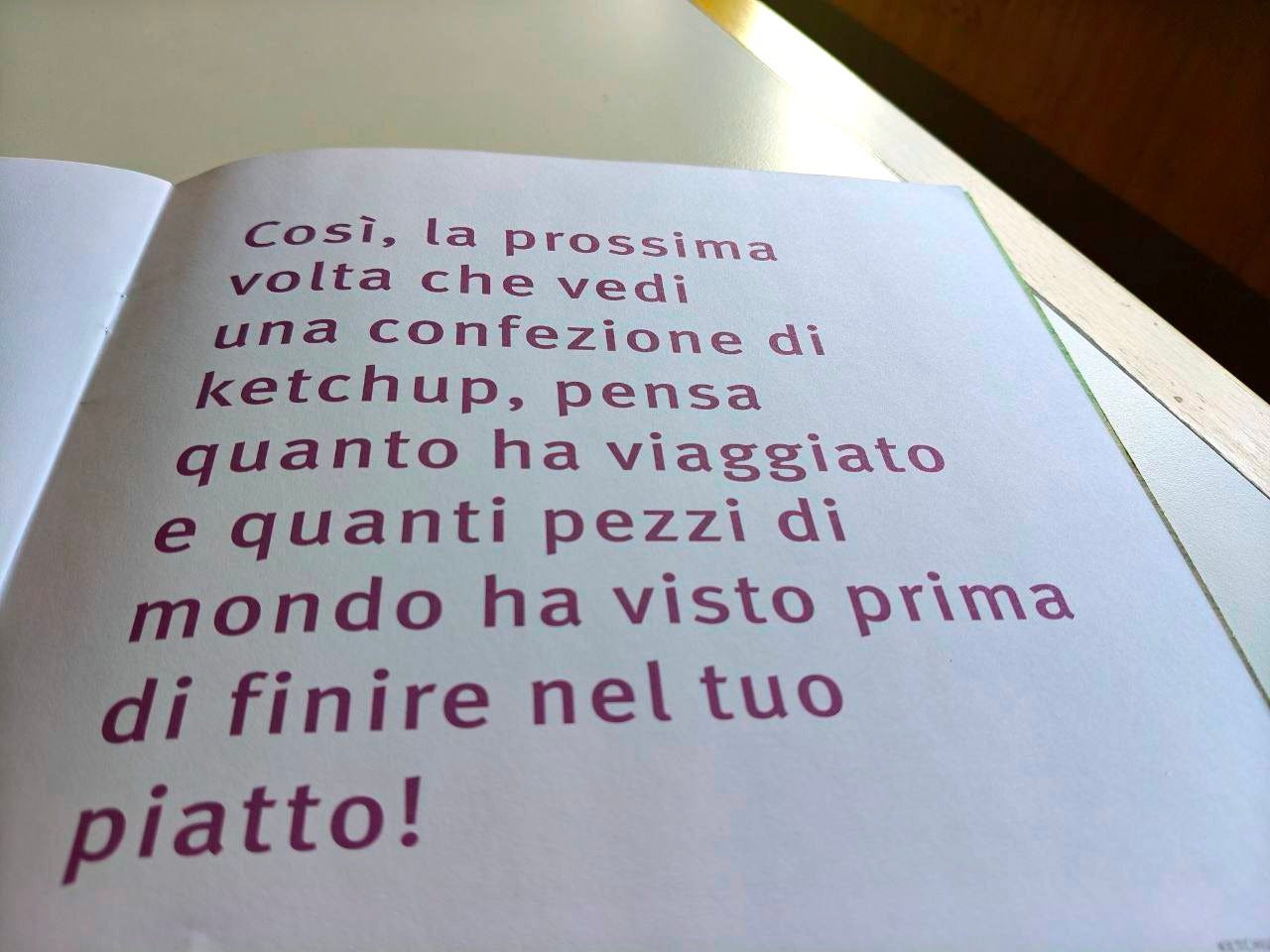 Una pagina della parola KETCHUP delle Scatoline di effequ, con questa scritta: "Così, la prossima volta che vedi una confezione di ketchup, pensa a quanto ha viaggiato e quanti posti nel mondo ha visto prima di finire nel tuo piatto". La pagina è appoggiata su un tavolo di legno bianco sporco e nell'angolino in alto a destra si intravede un pezzetto di muro in ombra.
