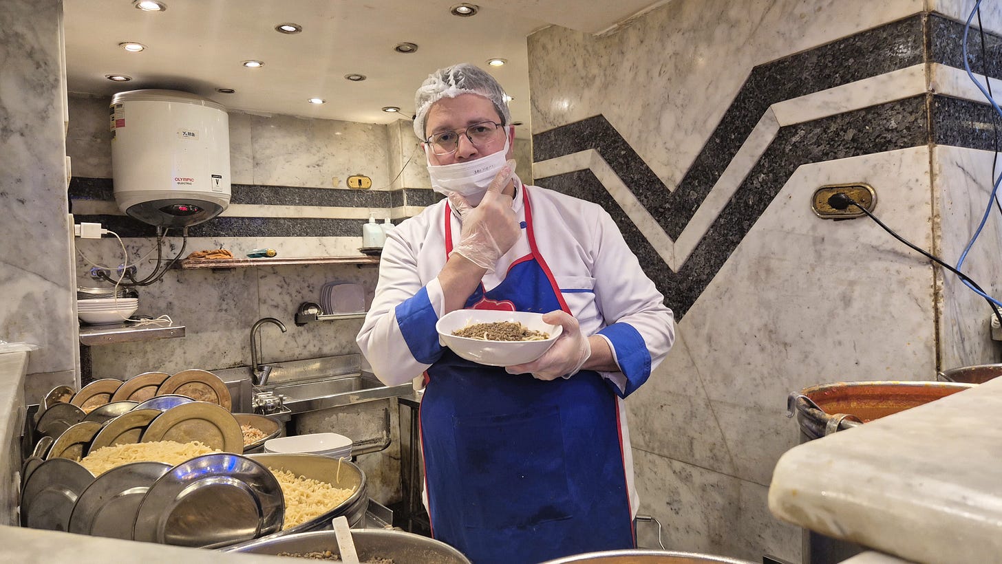 A man wearing an apron and a white face mask holds up a bowl of Koshary and looks toward the camera standing in a kitchen prep area