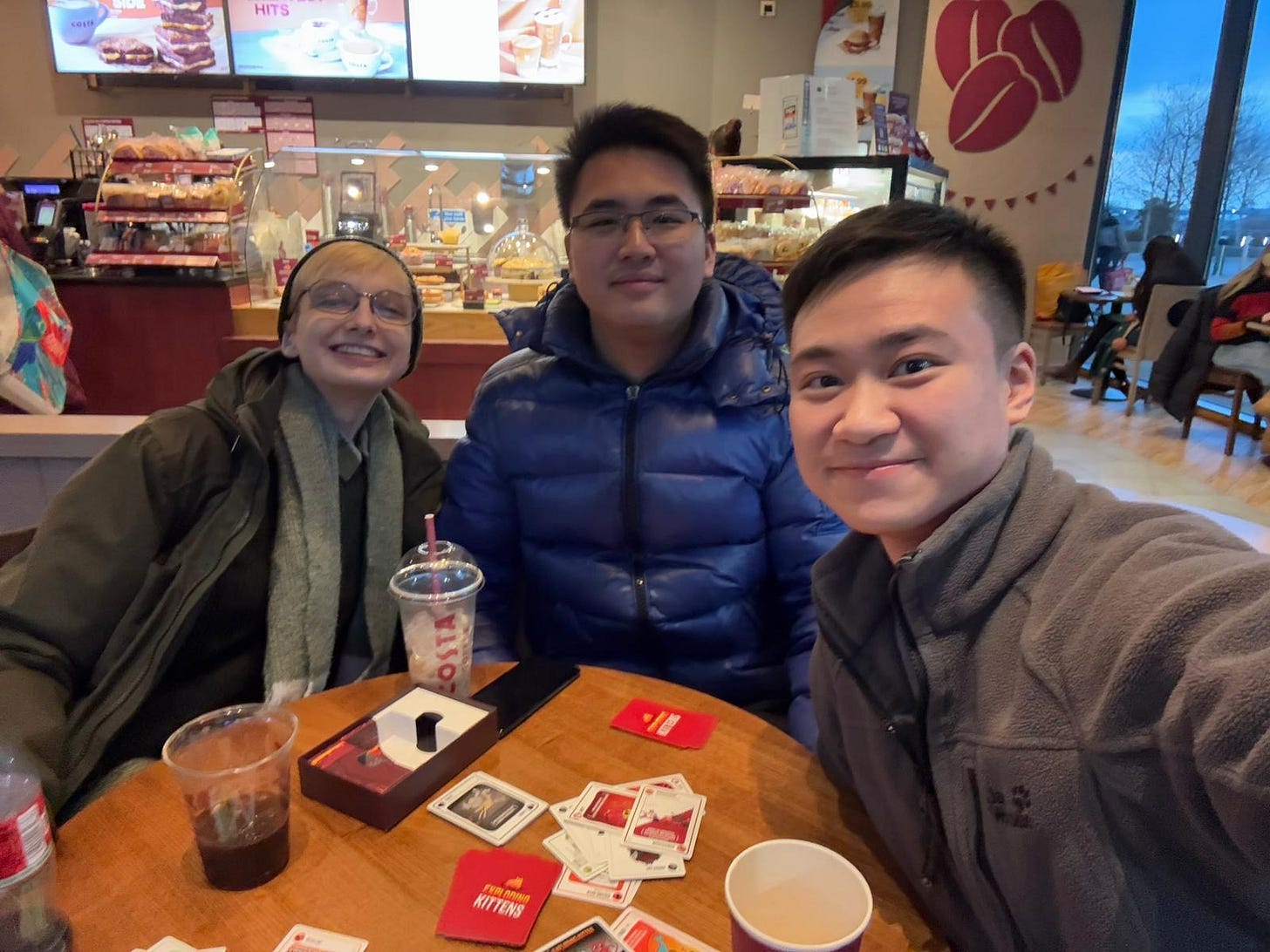 Beck, Shen and Aiden sitting in Costa playing the card game Exploding Kittens, smiling at the camera