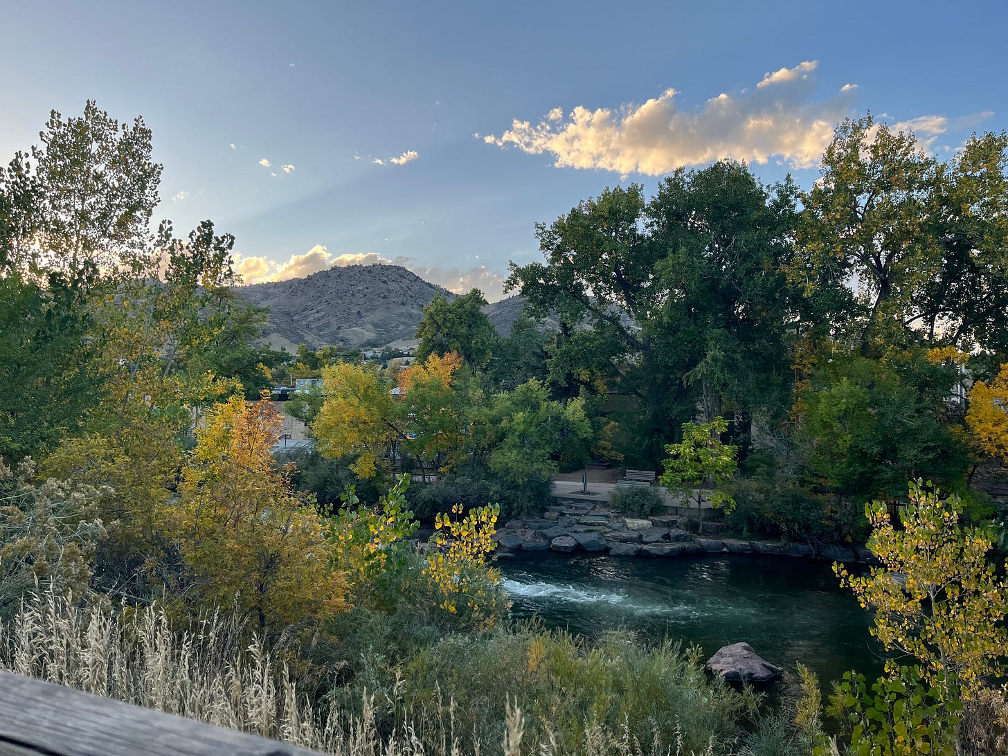 a photo of Clear Creek in Golden, CO with trees with green and yellow leaves, blue green water, and puffy white clouds