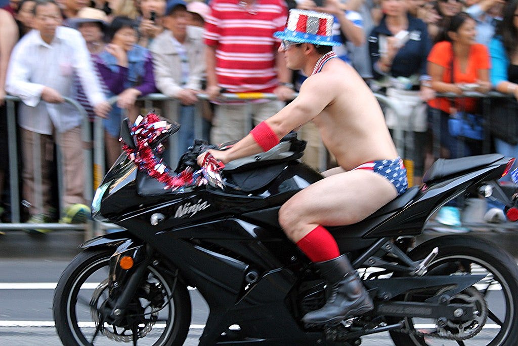 Image of a scantily-clad man on a black crotch-rocket-style motorcycle, wearing a red-white-and-blue Uncle Sam-style top hat with seqins and US-flag-themed briefs and high black boots, riding in what appears to be a parade, with onlookers on the side of the street. The man’s attire and bearing suggests a robust self-confidence.
