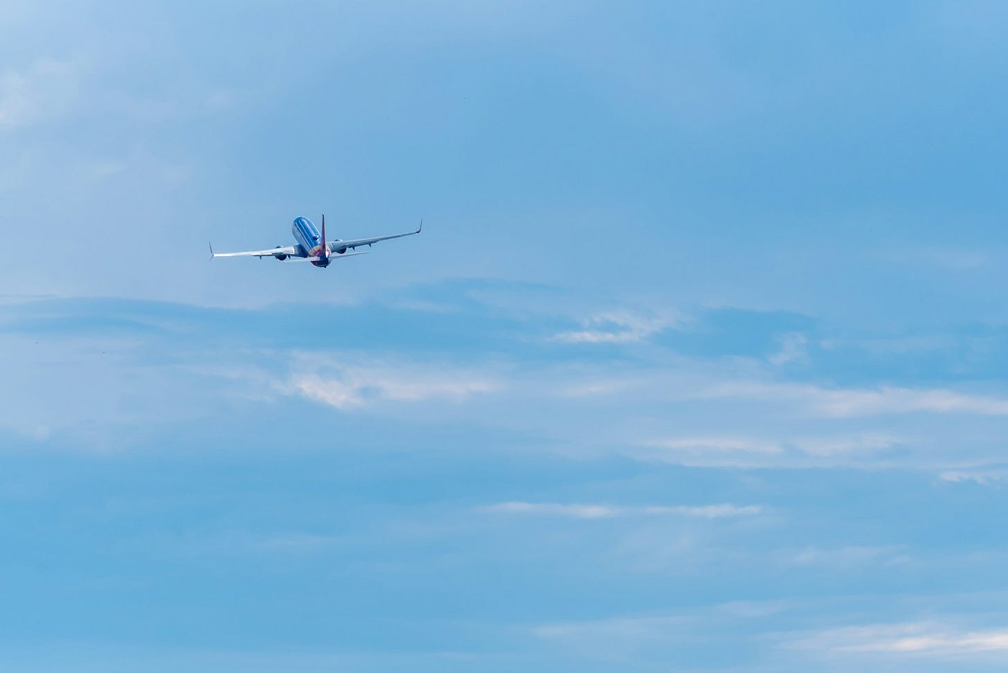 The image shows a commercial passenger aircraft in flight against a clear blue sky with some thin, wispy clouds. The plane appears to be ascending after takeoff, with its distinctive blue and white livery visible. It's captured from below, displaying the aircraft's underside as it flies against the bright daytime sky.
