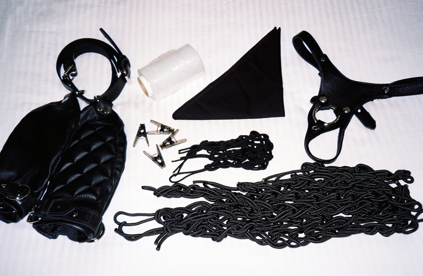 A flash film photo of a collection of items laid out on a white bed: padded black leather mitts with straps, four black clothespins, a roll of clear plastic wrap, a black bandana, piles of thin and thick black rope, and a black leather harness.