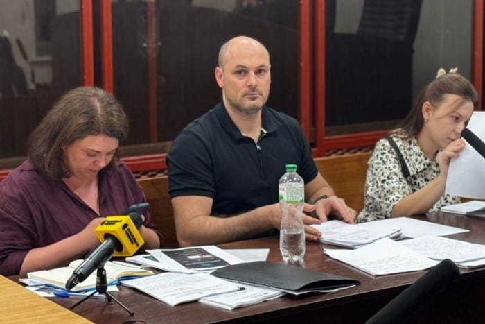 Ruslan Mahamedrasulov sits at a courtroom table with two women, surrounded by documents and a water bottle.