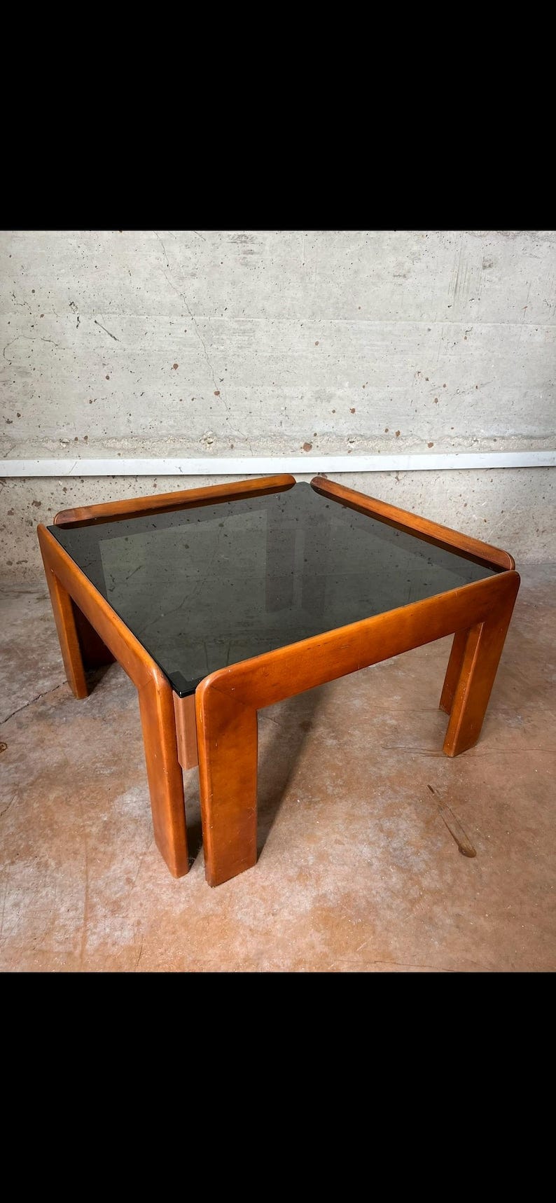 May include: A square coffee table with a dark glass top and a wooden frame. The frame is a light brown color and has a unique design with curved edges. The table is sitting on a concrete floor.