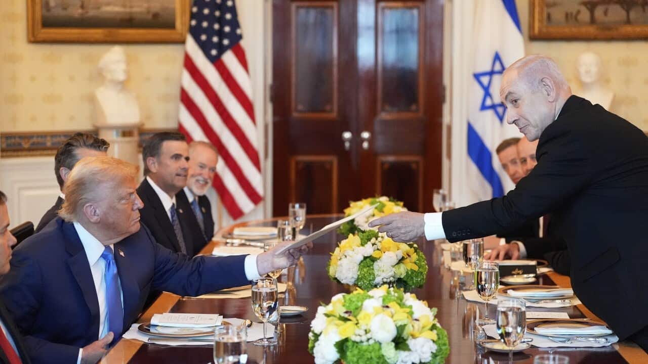 Donald Trump being handed an envelope by Benjamin Netanyahu at a dinner table, with other people sitting on either side. There are also American and Israeli flags.