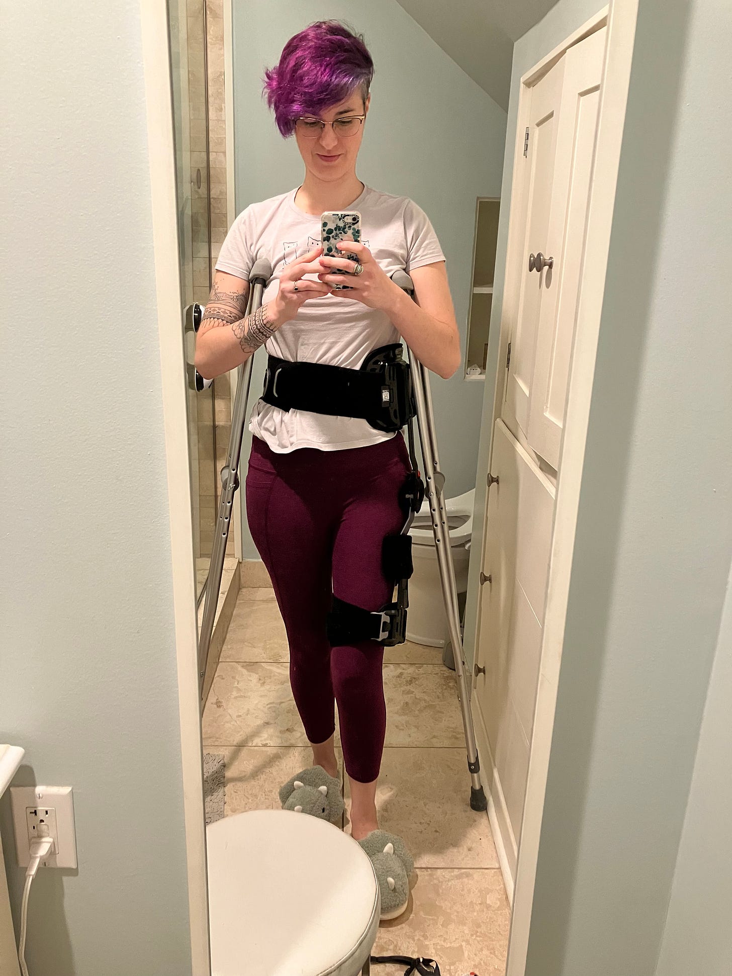 ALT Text: A white woman with short purple hair and glasses, looking down at her phone to take a mirror selfie. She's wearing a white shirt and purple leggings and propped up on crutches. Around her waist and extending down one leg to the knee is a large black brace. ALT Text: A white woman with short purple hair and glasses, looking down at her phone to take a mirror selfie. She's wearing a white shirt and purple leggings and propped up on crutches. Around her waist and extending down one leg to the knee is a large black brace.