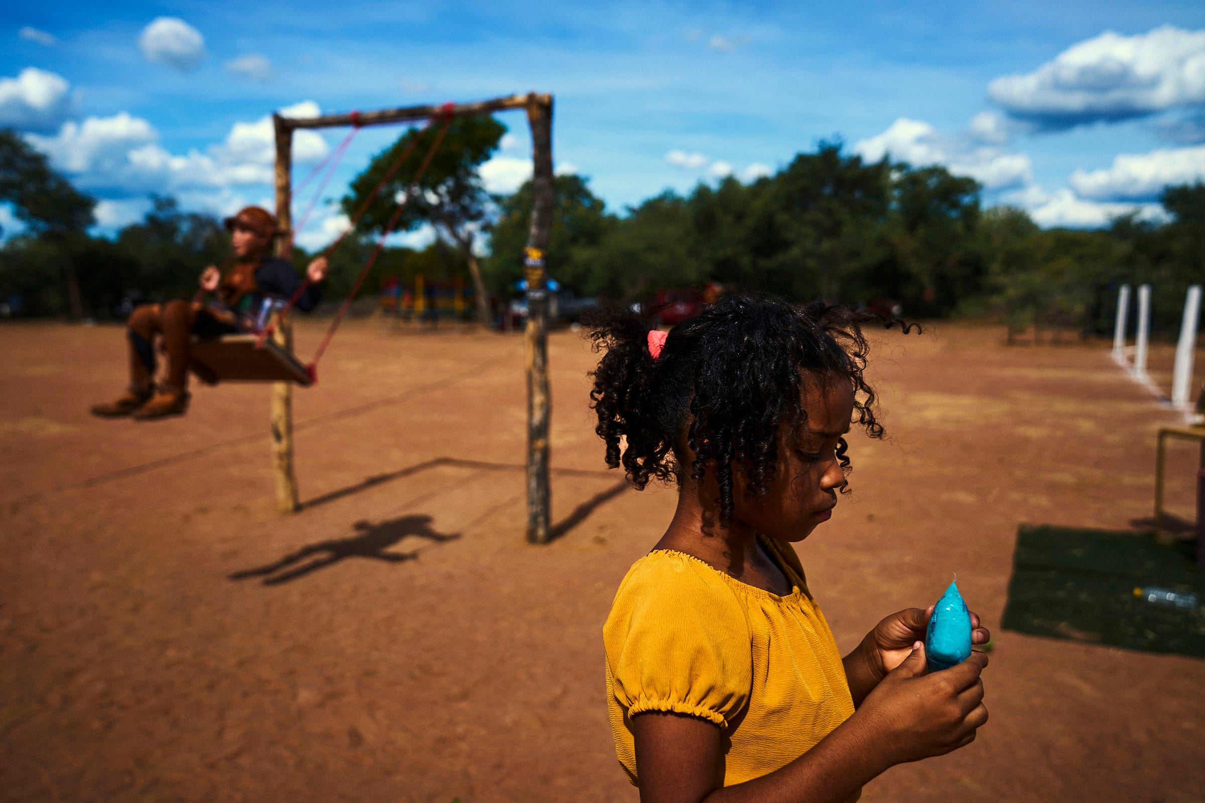 A young girl in a yellow shirt examines a blue balloon outdoors, while another child swings on a wooden swing in the blurred background under a partly cloudy sky.