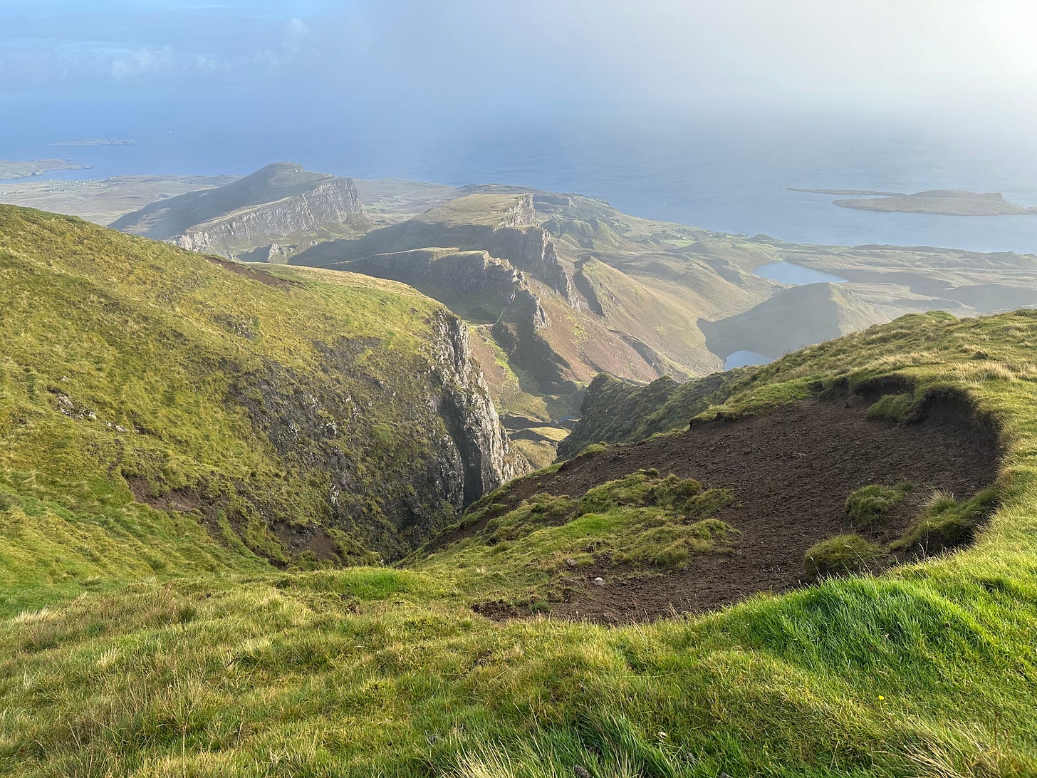 A view from up high on a very green mountain top, with lots of smaller green mountain cliffsides below and the blue ocean and sky beyond