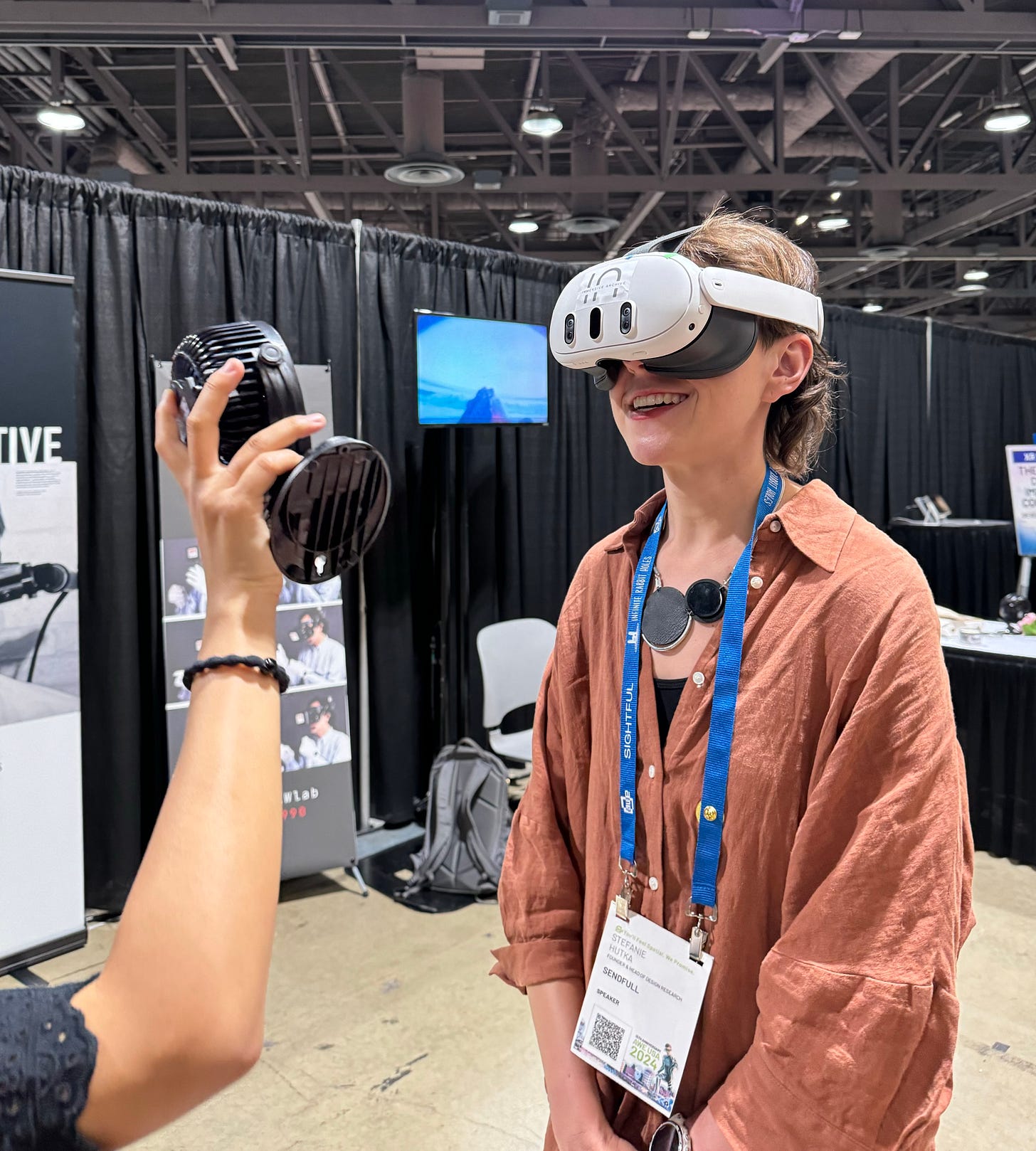 Stef, in a long brown shirt, wears a Quest 3 headset while another person holds a small fan up to her face. Stef, in a long brown shirt, wears a Quest 3 headset while another person holds a small fan up to her face.