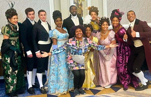A group of young actors all in colorful Regency costumes, gathered together and smiling, with executive producer Tia A. Smith in the foreground holding out her hands in celebration A group of young actors all in colorful Regency costumes, gathered together and smiling, with executive producer Tia A. Smith in the foreground holding out her hands in celebration