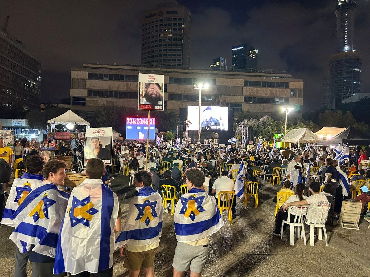 Nighttime outdoor square with crowds of people mostly men and women in casual clothing seated on plastic chairs under white tents. Israeli flags in blue and white with Star of David are draped over backs of chairs and worn by individuals. Large digital screen displays numbers 124 and 251 possibly countdown or identifiers. Giant poster of a bearded man on building facade. Background features tall modern skyscrapers lit up city lights and floodlights illuminating the area. Tents and banners set up for event setup.