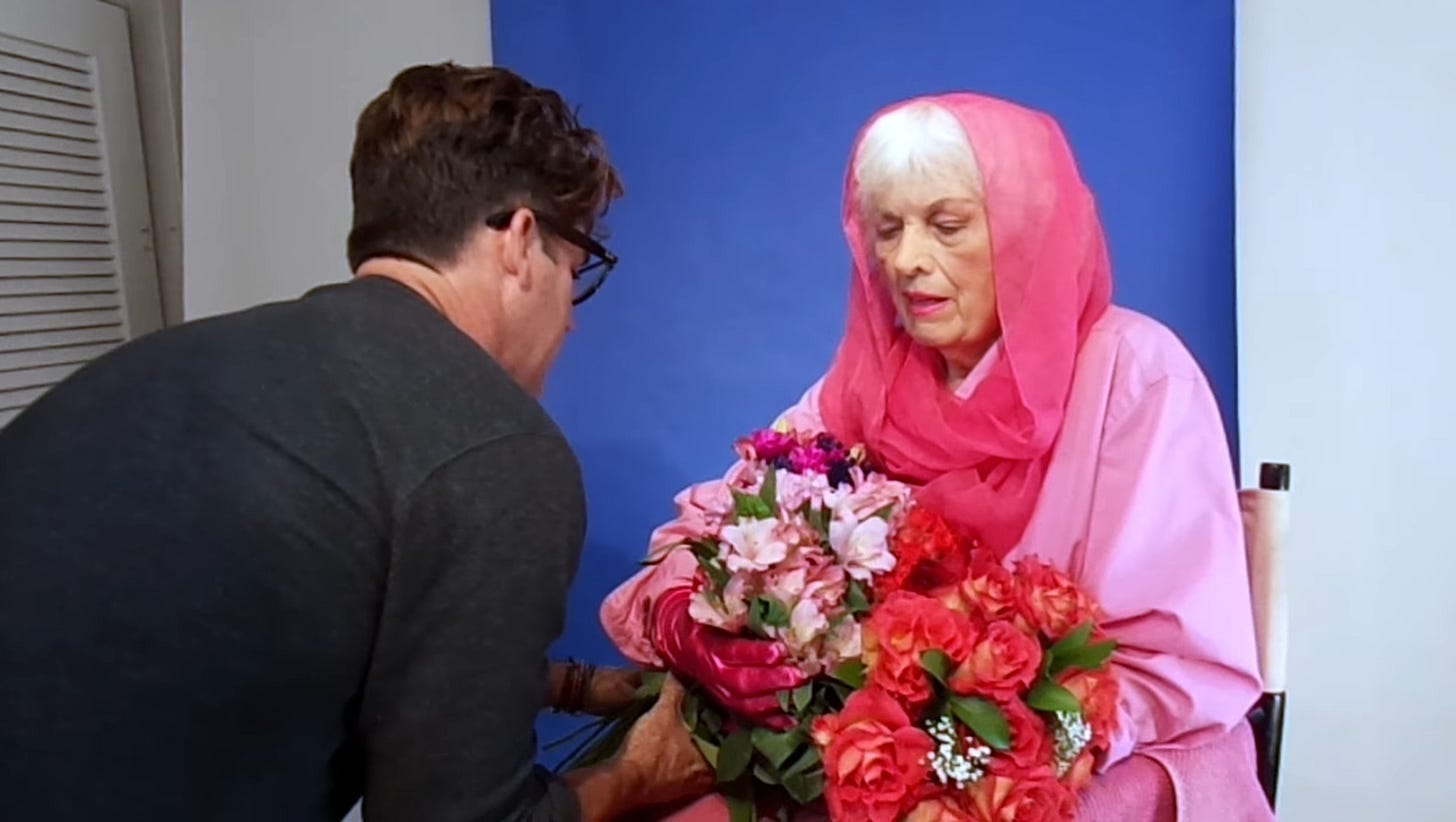 An elderly Bunny Yeager wearing a bright pink headscarf and gloves sits against a blue backdrop while holding two large flower bouquets, as a man in glasses adjusts the arrangement in front of her.