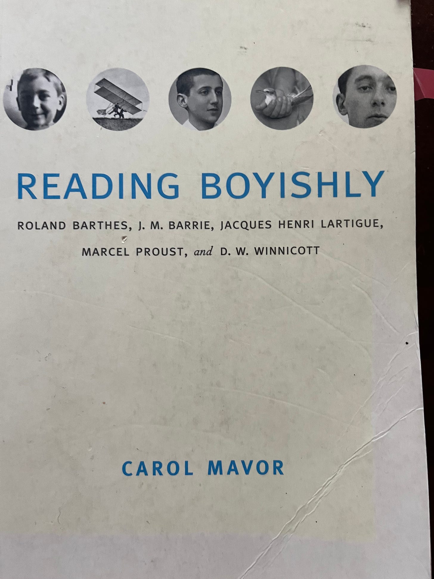 A scuffed cover of Reading Boyishly: Roland Barthes, J.M. Barrie, Jacques Henri Lartigue, Marcel Proust, and D. W. Winnicott. The title is in blue, and there circular pictures above this, with a child's face, a photo of a biplane, another child's face, a hand holding a crab claw, and still another child's face.