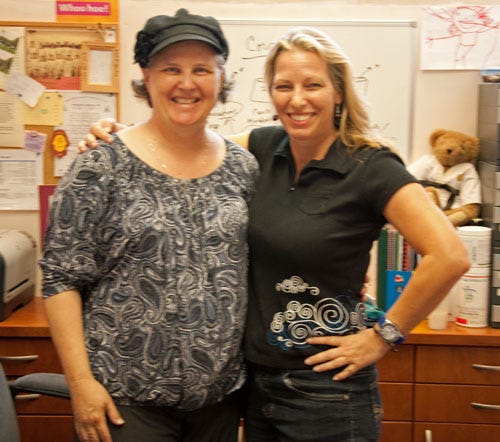 Two white women stand next to each other smiling and hugging. One is wearing a navy hat, the other is wearing a black polo shirt. 