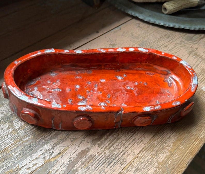 May include: An orange ceramic oval dish with white speckles and a raised rim. The rim has a row of evenly spaced, round, orange buttons.
