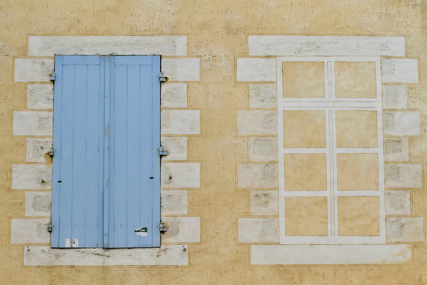 Parete esterna di un edificio di color giallino. Vi compaiono due finestre: a destra una finestra disegnata in bianco, con lo sfondo giallino del muro; a sinistra una finestra chiusa da imposte di legno color azzurrino. Parete esterna di un edificio di color giallino. Vi compaiono due finestre: a destra una finestra disegnata in bianco, con lo sfondo giallino del muro; a sinistra una finestra chiusa da imposte di legno color azzurrino.
