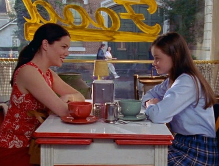 This may contain: two women sitting at a table talking to each other in front of a food sign