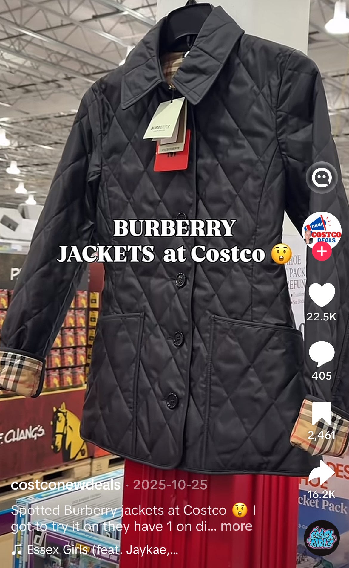 TikTok screenshot from creator @costconewdeals dated October 25, 2025, documenting an authentic Burberry women's black quilted diamond-stitch jacket being sold at a US Costco Wholesale warehouse store. The image shows the jacket hanging on display with the iconic Burberry check pattern lining clearly visible at the folded cuffs (the signature camel, black, white, and red tartan check) revealing the brand's heritage interior detailing. The jacket features classic diamond quilting across the body, a structured pointed collar, two front patch pockets, and black tonal buttons including a logo-engraved Burberry button at the collar closure. Multiple Burberry hangtags in cream and signature Burberry red are clearly visible at the neckline, removing any ambiguity about brand authenticity. The display is positioned in front of a Costco merchandise endcap stocked with branded food and household goods, with the warehouse's characteristic fluorescent lighting and exposed industrial ceiling visible in the background. Bold white serif text overlay reads "BURBERRY JACKETS at Costco" with a surprised face emoji. The TikTok post caption reads "Spotted Burberry jackets at Costco. I got to try it on they have 1 on display," and the video has accumulated 22,500 likes, 405 comments, 2,461 saves, and 16,200 shares (one of the highest-engagement Burberry-at-Costco posts on TikTok). This is the Burberry diamond-quilted jacket silhouette, a long-established core outerwear style in the brand's heritage archive that the company has cited as central to its current heritage refocus and pricing-by-authority strategy. Channel-check evidence material to Burberry $BRBY.L investors evaluating brand positioning discipline, parallel-channel inventory clearance, and the credibility of management's articulated turnaround thesis under CEO Joshua Schulman, particularly given that diamond-quilted outerwear sits in the exact "hero category" the company says is anchoring its return to authority-based pricing. Notable that on a recent Intern Pierre primary research call, a former member of the Office of the CEO with 2.5 years of tenure stated they had never heard of Burberry being sold at Costco, despite this single post reaching over 16,000 shares.