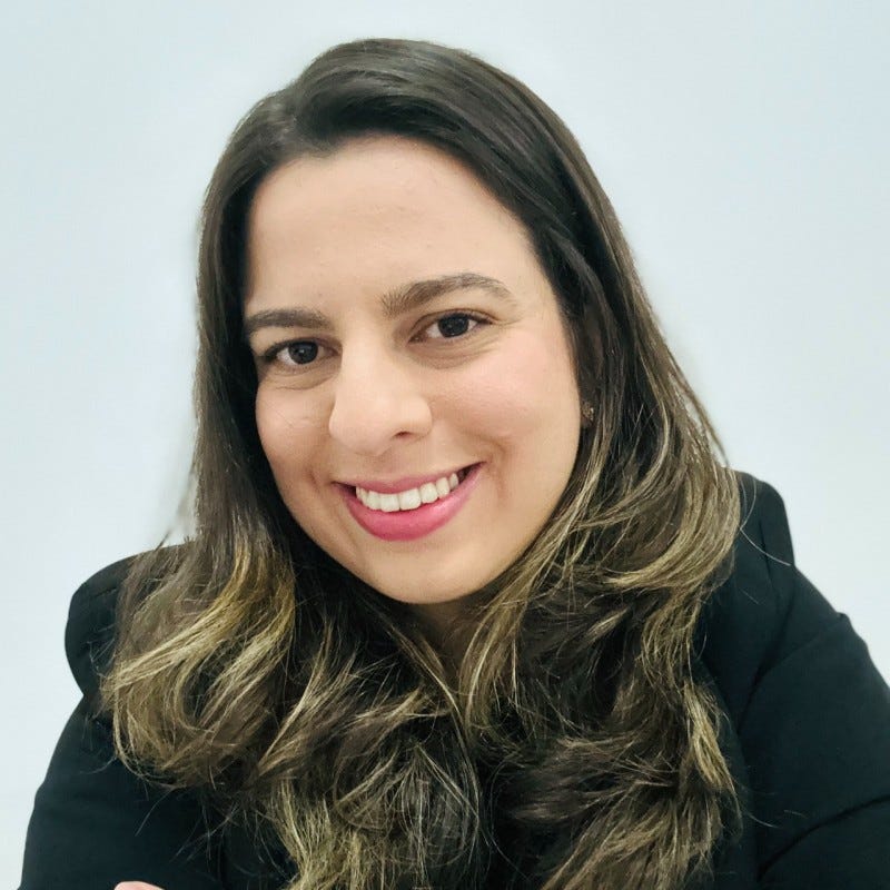 A imagem contém uma mulher sorrindo, com cabelos castanhos longos e ondulados com luzes. Ela usa batom rosa escuro e um paletó preto formal. O fundo é branco. A imagem contém uma mulher sorrindo, com cabelos castanhos longos e ondulados com luzes. Ela usa batom rosa escuro e um paletó preto formal. O fundo é branco.