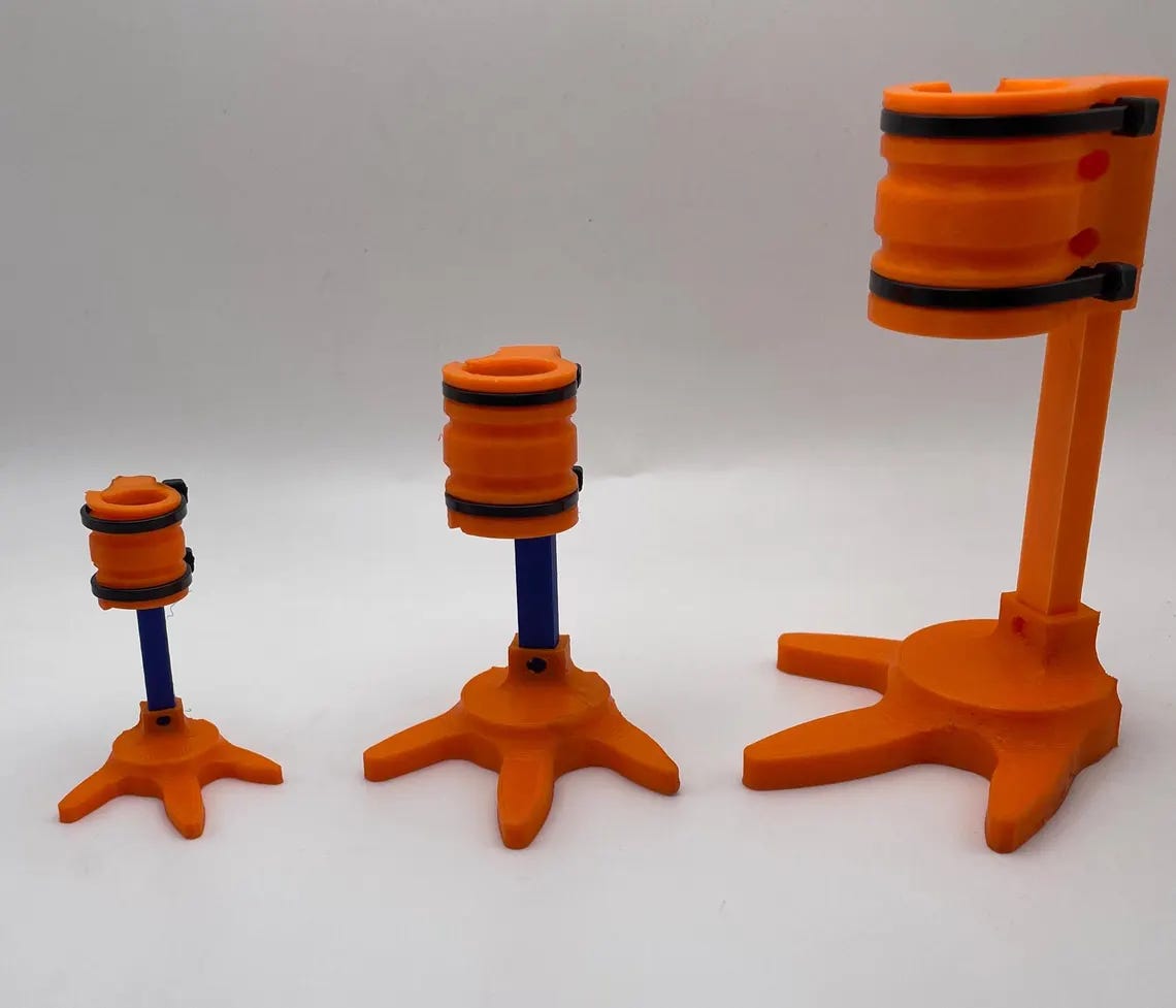 Three 3D-printed prosthetic chicken legs in small, medium, and large sizes. Each has an orange base with a three-pronged foot and a cylindrical orange leg socket held by a vertical blue rod. The devices are designed for chickens and vary in height and thickness.