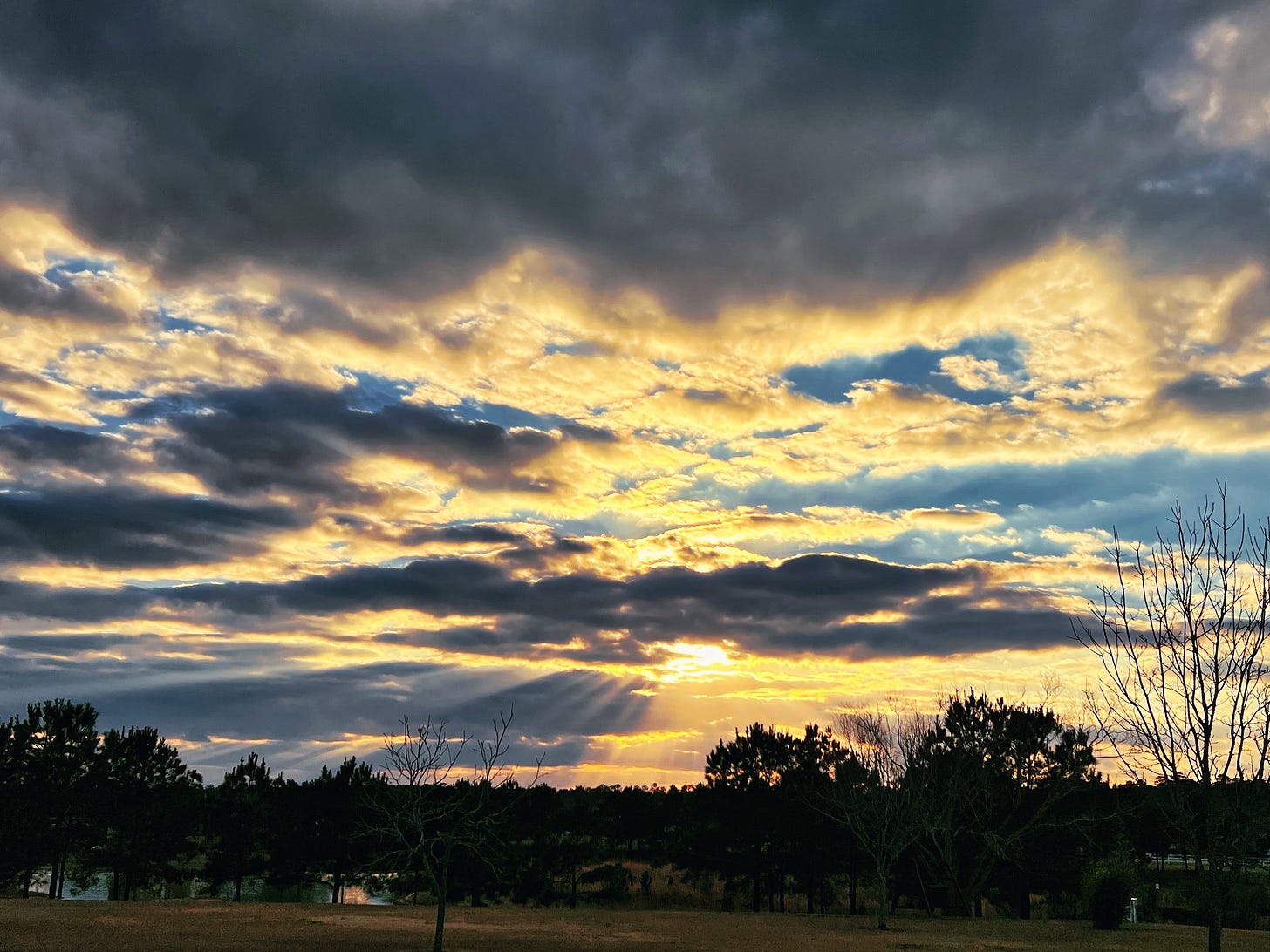 The sun peers out behind the blue-grey and yellowish clouds against a blue sky and a lake partially hidden behind pine trees at the bottom of the photo.