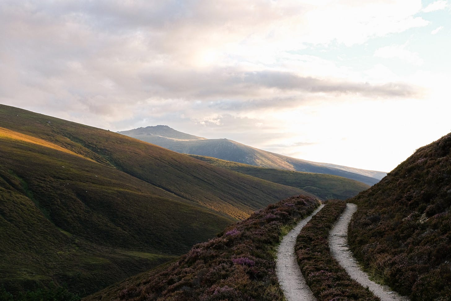 A Landover track winds its way through think purple heather along a steel hillside. In the back ground towering hills merge with the sky creating a sense of scale. 
