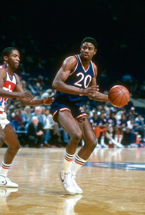 Michael Ray Richardson is seen dribbling the ball with his back to a defender. He wears a blue Knicks uniform.