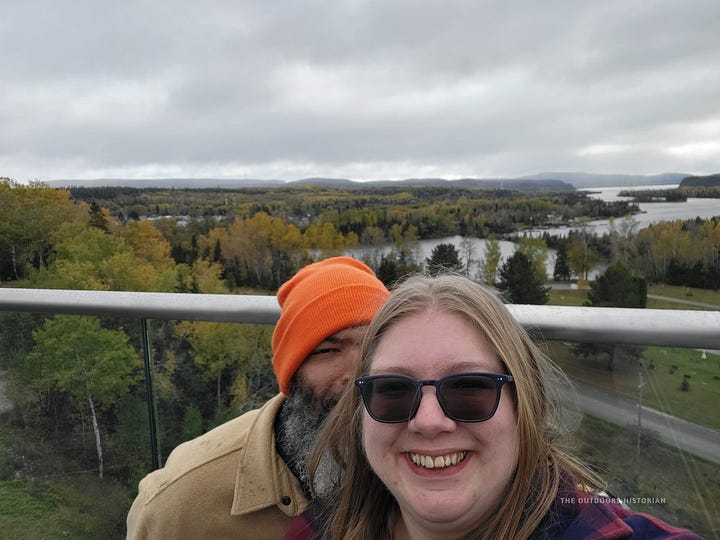 Image 1: Eugene smiling and holding up two takeaway coffee cups in front of the glass windows of a building. Image 2: A selfie of Emily and Eugene on a windy overlook, wearing jackets, with a grey sky and yellow and green forest behind them.