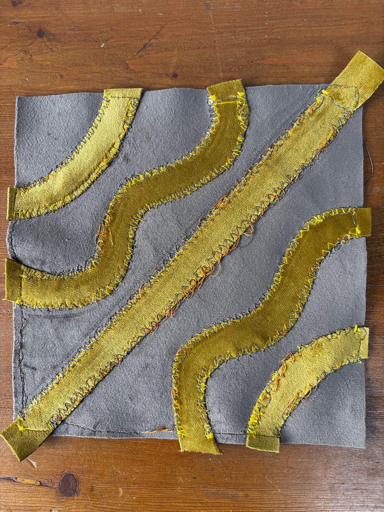 A 20 centimetre by 20 centimetre square of grey velvet on a wood surface. The grey velvet has 2 centimetre patterns of yellow material sewn onto it. The sewing in places shows very loose orange thread.