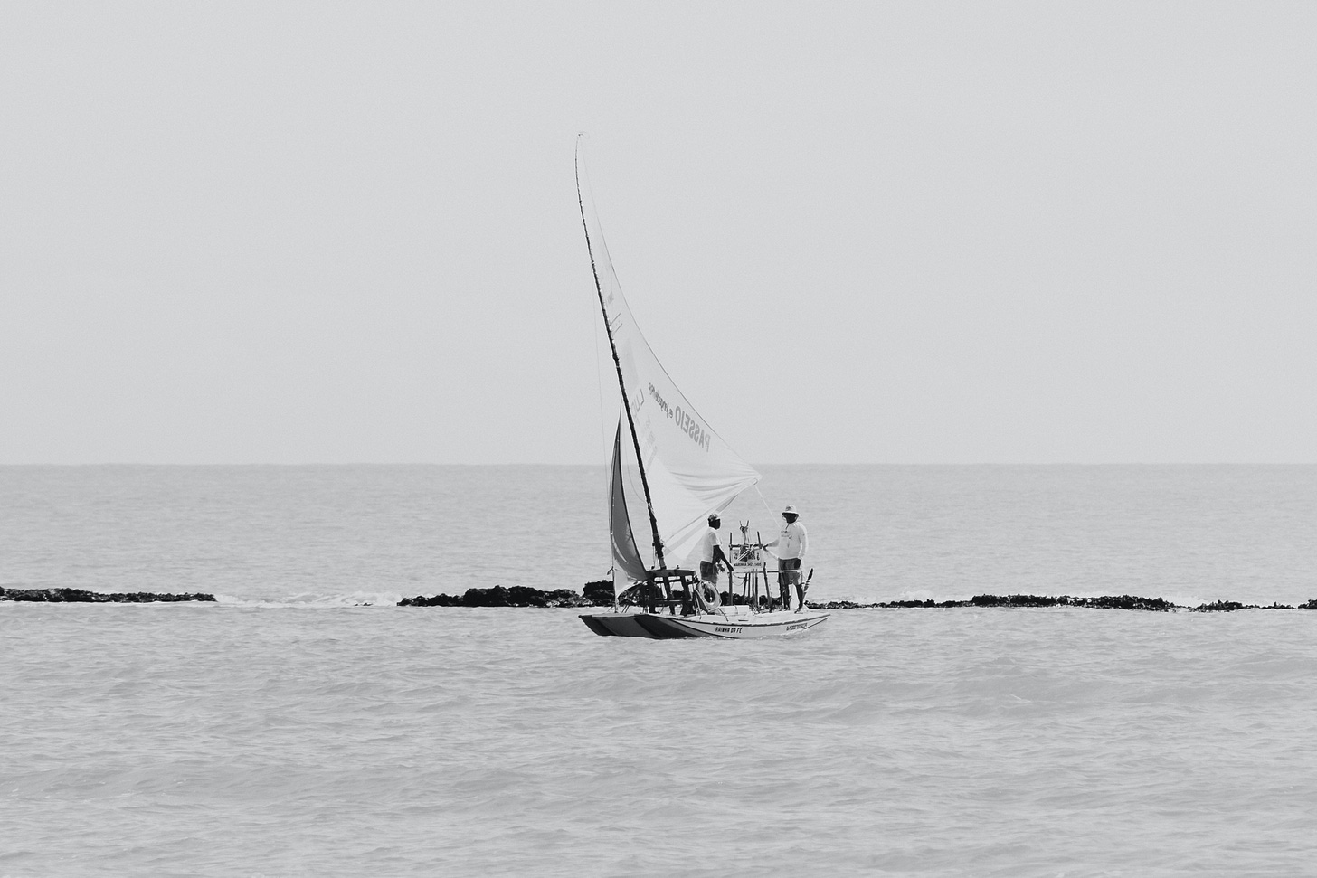 Imagem em preto e branco. No centro da foto, dois homens de pé sobre uma jangada a vela. A jangada está próxima de uma barreira de corais. Toda a metade inferior da imagem mostra o mar, e a metade superior, o céu