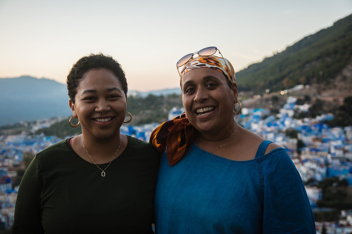 Anela stands with a woman, both smiling at the camera, with blue and white dotted buildings behind them.