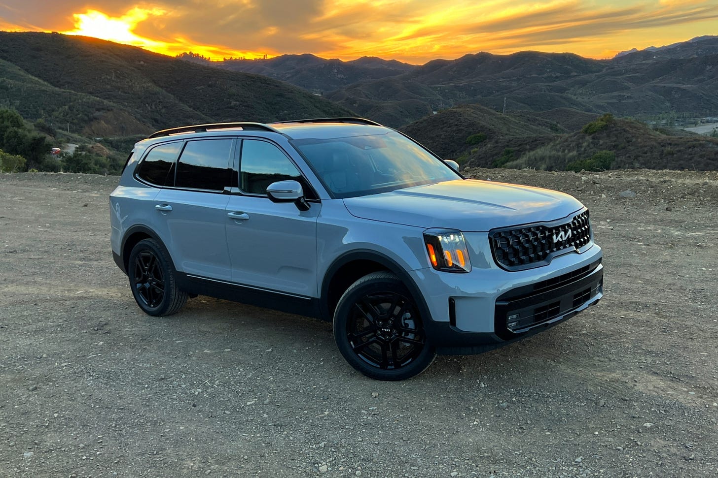 A gray 2024 Kia Telluride is parked on gravel near mountains at sunset.