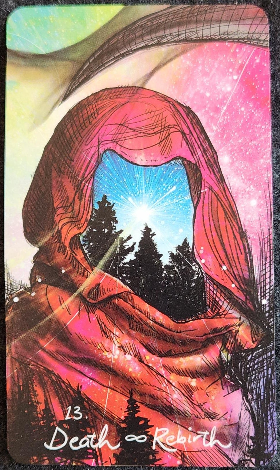 A tarot card labelled 13. Death ∞ Rebirth. The card shows a hooded figure, but through the hood instead of a face there is a row of trees and a dark blue sky with a burst of light. The hood is a deep red. In front of the hooded figure are more trees. The sky behind is a mix of colours, pink and green and speckled in stars.