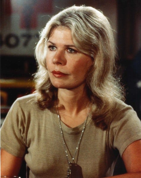 Margaret Houlihan, woman in khaki army t-shirt and dog-tags, blond medium-length hair, red lipstick, looking out into the distance with a look of determination