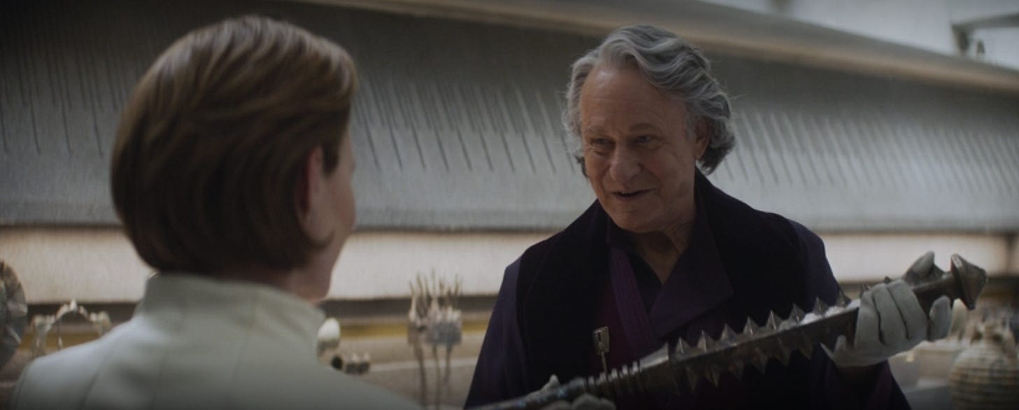 We see the back of a woman's head and an older man, dressed flamboyantly as an art dealer, presents her an antique.