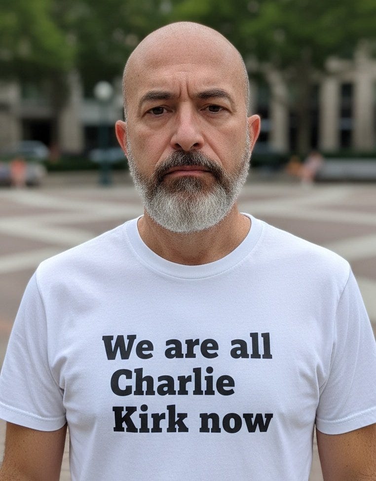 A man with a bald head and gray beard wearing a white T-shirt with the text "We are all Charlie Kirk now" in black. The background shows an outdoor setting with trees, a paved area, and buildings.