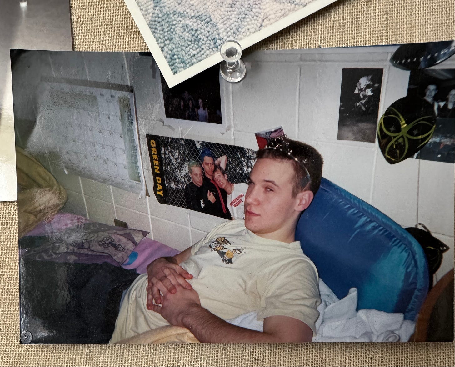 a photograph on a pinboard of a young white man reclining in a dorm room, hands folded on his stomach, wearing a tiara a photograph on a pinboard of a young white man reclining in a dorm room, hands folded on his stomach, wearing a tiara