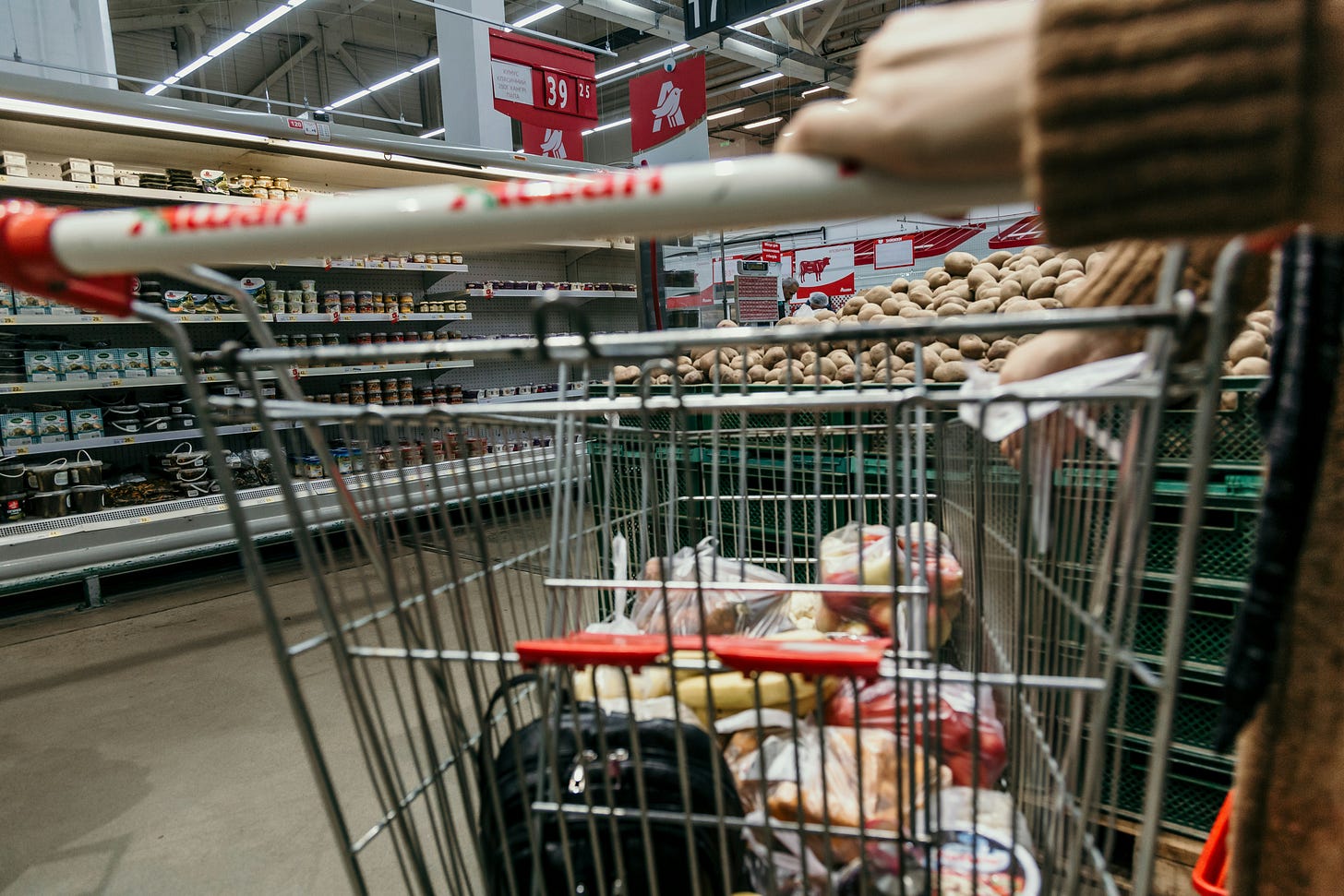 A shopping trolley with groceries in it being pushed by a woman in a brown cardigan.