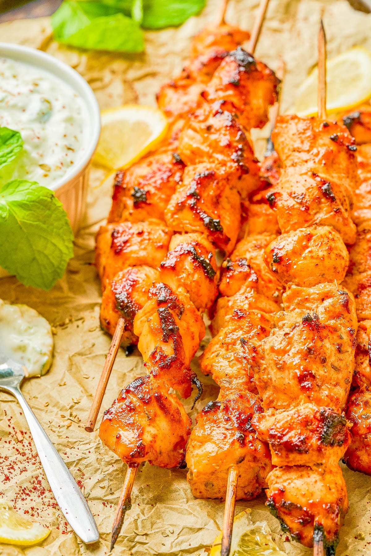 Grilled chicken skewers with a reddish marinade are arranged on parchment paper, accompanied by a bowl of yogurt sauce, mint leaves, and lemon slices.