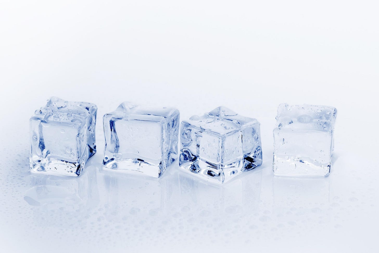 Four clear ice cubes on a white surface, glistening with droplets against a bright minimal background. Four clear ice cubes on a white surface, glistening with droplets against a bright minimal background.