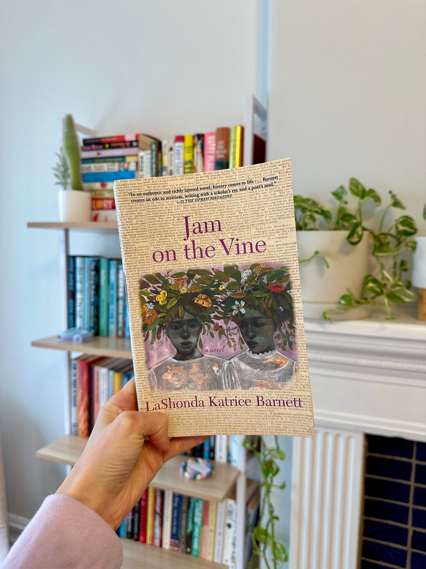 A white hand holds up a copy of Jam on the Vine in front of a fireplace and bookshelf. The book has a cover with newspaper print and two individuals wearing floral headpieces.