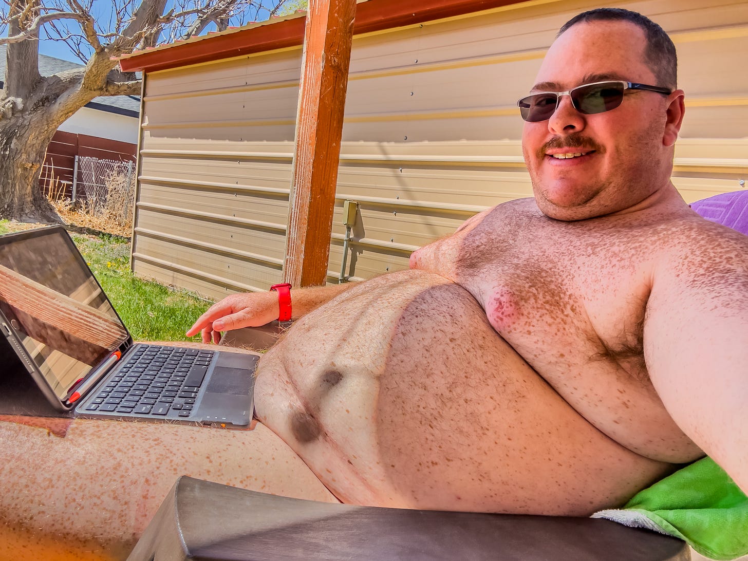 A close-up, outdoor photo of the author seated in a chair completely nude, working on a laptop while relaxing under a covered patio on a sunny day. The man is smiling at the camera, wearing dark sunglasses, and is shirtless, with a visible wristwatch on his left wrist. A small, black laptop (or tablet with a keyboard cover) rests on his lap. In the background, there is a corrugated metal fence or wall, a grassy yard, and a large tree trunk to the left. A wooden support post for the patio cover is visible directly next to the man on the left.