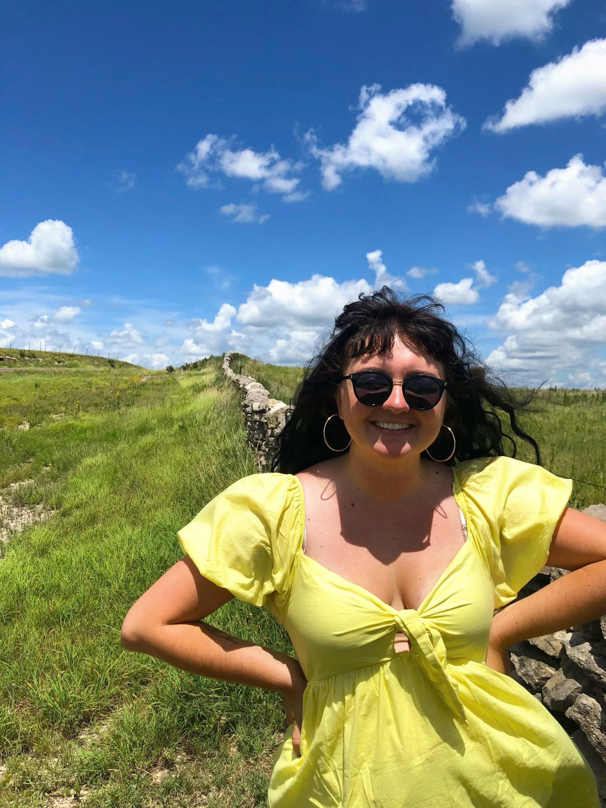 A smiling woman in a bright yellow dress with puff sleeves stands outdoors on a sunny day, wearing sunglasses and hoop earrings. Behind her is a grassy hill with a stone wall winding into the distance under a vivid blue sky with scattered clouds.