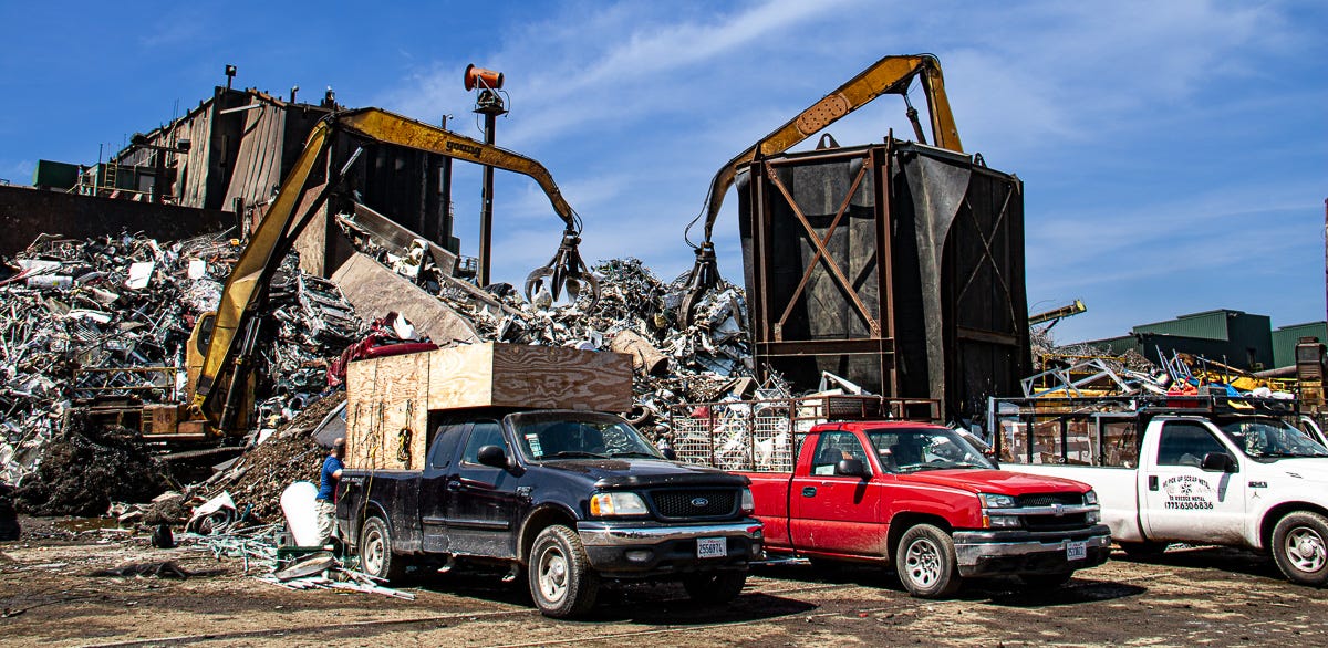 Multiple pickup trucks, some heavily modified, parked in front of active cranes and towering scrap piles inside the yard.